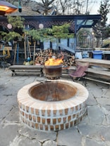 A cozy outdoor setting featuring a fire pit in the center, surrounded by a stone patio. There is a stack of firewood next to a wooden structure adorned with string lights and greenery. A chair is placed nearby, creating a warm and inviting atmosphere.