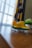 A small toy airplane with a bright yellow and green color scheme sits on a wooden surface near a window. The background features a geometric-patterned curtain in shades of gray and yellow, with a blurred view of a potted plant nearby.
