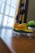A small toy airplane with a bright yellow and green color scheme sits on a wooden surface near a window. The background features a geometric-patterned curtain in shades of gray and yellow, with a blurred view of a potted plant nearby.