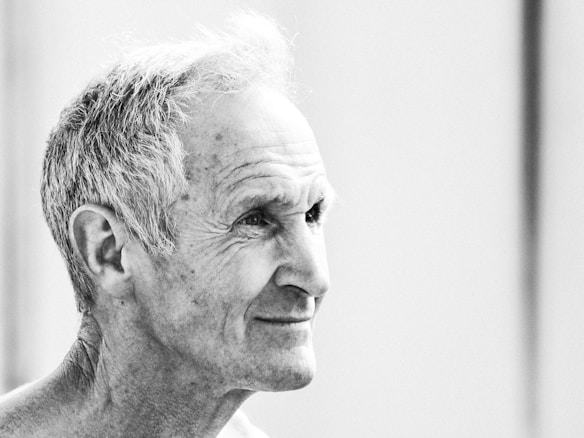 A black and white portrait of an older man with short, grey hair. His face is turned in profile, revealing a gentle smile and features highlighted by subtle shadows. Wrinkles are visible, suggesting experience and wisdom.
