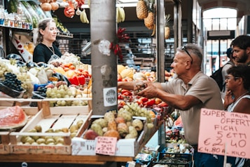 Vibrant market scene with a variety of fresh fruits and vegetables displayed at a stall. A vendor, wearing a black shirt, is attending to a customer who is selecting produce. The stall is filled with grapes, melons, pineapples, and other colorful fruits. There are several people in the market, including a man with gray hair and sunglasses resting on his head, engaging with the produce. Signs with prices and offers are visible on the stalls.