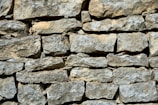 A wall constructed from irregularly shaped stones, stacked together without visible mortar. The surface of the stones is rough and textured, with a mix of light gray and beige colors. The arrangement gives a rustic and natural appearance.