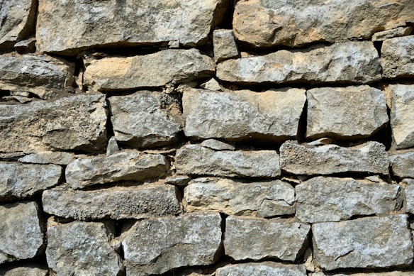 A wall constructed from irregularly shaped stones, stacked together without visible mortar. The surface of the stones is rough and textured, with a mix of light gray and beige colors. The arrangement gives a rustic and natural appearance.