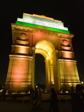 A large, illuminated monument stands tall against a dark night sky. The structure is lit in vibrant colors of orange, white, and green, resembling the Indian national flag. People are visible at the base, walking and observing the monument, which has an arched passageway and architectural detailing.