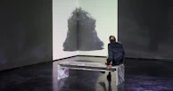 Visitors seated on minimalist benches, deeply absorbed in a multimedia queer art exhibit projected on the gallery wall.