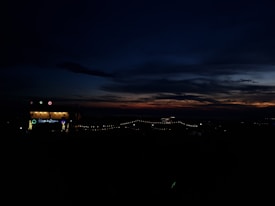 A dark outdoor scene with a vivid sunset casting shades of orange and purple across the sky. There are string lights illuminating a pathway, along with a lit sign and some decorative lights in the left part of the image, indicating some kind of event or place.