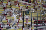 Various colorful and intricate hanging ornaments are set against a patterned striped wall. The ornaments feature designs such as concentric circles, floral shapes, and geometric patterns, some incorporating mirrored and metallic textures.