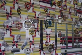 Various colorful and intricate hanging ornaments are set against a patterned striped wall. The ornaments feature designs such as concentric circles, floral shapes, and geometric patterns, some incorporating mirrored and metallic textures.