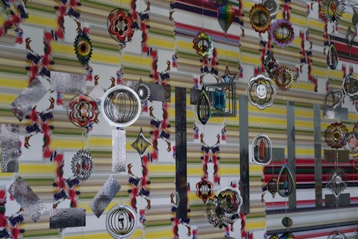 Various colorful and intricate hanging ornaments are set against a patterned striped wall. The ornaments feature designs such as concentric circles, floral shapes, and geometric patterns, some incorporating mirrored and metallic textures.