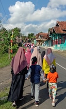 A group of people, including women wearing hijabs and children in casual clothing, are walking down a rural road lined with colorful houses and greenery. The sky is bright with large fluffy clouds, suggesting a clear and sunny day.