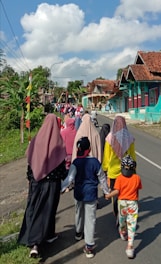 A group of people, including women wearing hijabs and children in casual clothing, are walking down a rural road lined with colorful houses and greenery. The sky is bright with large fluffy clouds, suggesting a clear and sunny day.