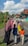 A group of people, including women wearing hijabs and children in casual clothing, are walking down a rural road lined with colorful houses and greenery. The sky is bright with large fluffy clouds, suggesting a clear and sunny day.