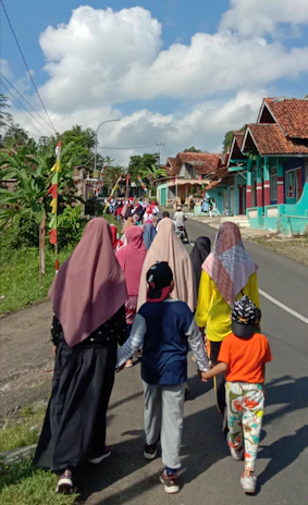 A group of people, including women wearing hijabs and children in casual clothing, are walking down a rural road lined with colorful houses and greenery. The sky is bright with large fluffy clouds, suggesting a clear and sunny day.
