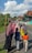 A group of people, including women wearing hijabs and children in casual clothing, are walking down a rural road lined with colorful houses and greenery. The sky is bright with large fluffy clouds, suggesting a clear and sunny day.