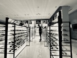 A monochrome view of a modern eyewear store featuring several illuminated display stands filled with sunglasses. A person stands in the aisle, likely browsing the collection. Advertising posters of various brands are visible on the walls above the displays.