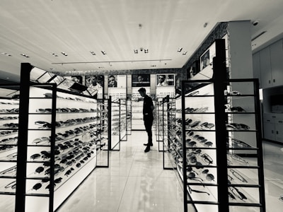 A monochrome view of a modern eyewear store featuring several illuminated display stands filled with sunglasses. A person stands in the aisle, likely browsing the collection. Advertising posters of various brands are visible on the walls above the displays.