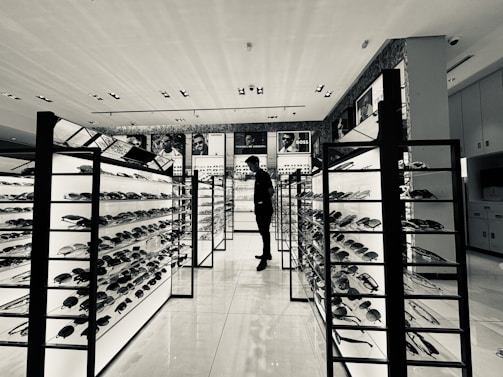 A monochrome view of a modern eyewear store featuring several illuminated display stands filled with sunglasses. A person stands in the aisle, likely browsing the collection. Advertising posters of various brands are visible on the walls above the displays.