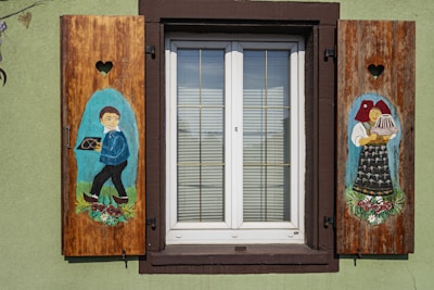 Charming window shutters feature vibrant painted figures. On the left, a boy in traditional attire carries a pretzel, while on the right, a woman in folk dress holds a cake. Both paintings are surrounded by floral designs, and heart-shaped cutouts adorn the shutters. The window is framed by a brown border, and green walls complete the quaint setting.