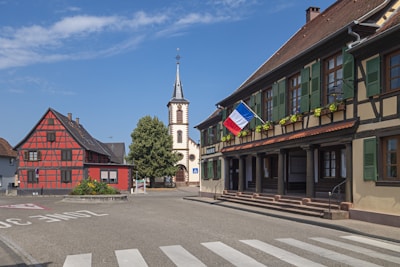 A quaint village scene with traditional half-timbered houses, one featuring a red and black design. A French flag is displayed prominently on a building with green shutters and potted flowers along the ledge. In the background, a church with a tall steeple stands next to a large green tree. The road in the foreground is marked as a 30 km/h zone.
