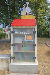 A small library box designed to resemble a red-roofed house is placed outdoors. The box features a colorful illustration of a stork holding a bundle in its beak on top of the structure. French text 'Votre boîte à livres' is displayed on the front, indicating a shared public book exchange. The box is set against a backdrop of trees and grass, suggesting a park or community area.