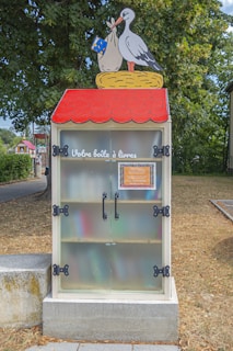 A small library box designed to resemble a red-roofed house is placed outdoors. The box features a colorful illustration of a stork holding a bundle in its beak on top of the structure. French text 'Votre boîte à livres' is displayed on the front, indicating a shared public book exchange. The box is set against a backdrop of trees and grass, suggesting a park or community area.