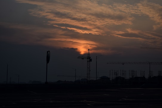The image depicts a sunset scene with a construction site in silhouette. The sky is filled with dramatic clouds that are illuminated by the warm glow of the setting sun. Several construction cranes are visible against the backdrop of the colorful sky.