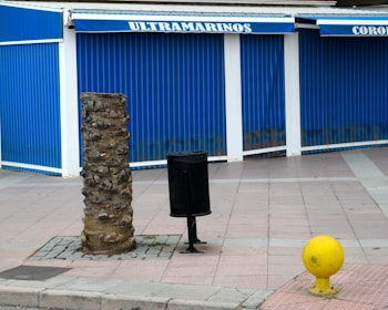 A blue store with closed shutters is situated in the background, and it has a sign reading 'ULTRAMARINOS.' In the foreground, there is a cut tree trunk, a black cylindrical trash bin, and a yellow spherical object with a post. The pavement is tiled with a pinkish hue.
