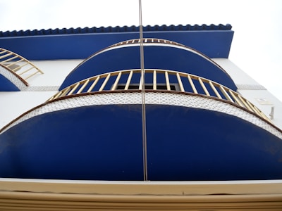 A modern architectural design featuring curved blue balconies with white grid-patterned bases and golden railings. The building has a clean, minimalistic style with a smooth white facade. The top is adorned with a blue corrugated roof.