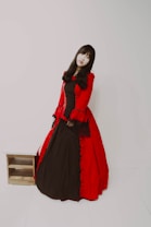 A person with long dark hair wearing a full-length gown featuring a deep red outer layer with black lace accents and a black underlayer. The outfit has a vintage, possibly gothic style. The background is plain and minimalistic, emphasizing the attire.