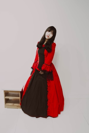 A person with long dark hair wearing a full-length gown featuring a deep red outer layer with black lace accents and a black underlayer. The outfit has a vintage, possibly gothic style. The background is plain and minimalistic, emphasizing the attire.