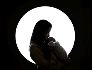 A silhouette of a person holding a baby is set against a bright circular light background. The person and child appear to be in a tender embrace, with soft lighting creating a peaceful and intimate atmosphere.