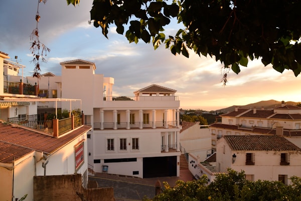 The image depicts a picturesque view of a Mediterranean-style residential area during sunset. The buildings have white facades with terracotta roofs, and balconies adorned with potted plants. The sky is filled with warm hues of orange and pink as the sun sets in the background.
