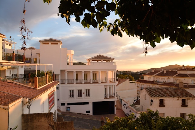 The image depicts a picturesque view of a Mediterranean-style residential area during sunset. The buildings have white facades with terracotta roofs, and balconies adorned with potted plants. The sky is filled with warm hues of orange and pink as the sun sets in the background.