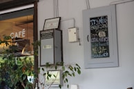 A cafe entrance features a glass door with the word 'Café' printed on it. To the right of the door is a chalkboard sign displaying a menu promoting a new cheese sourced from local harvests. The white wall also has a small no-smoking sign and some greenery, adding a touch of nature to the urban setting.