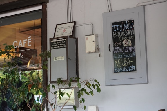 A cafe entrance features a glass door with the word 'Café' printed on it. To the right of the door is a chalkboard sign displaying a menu promoting a new cheese sourced from local harvests. The white wall also has a small no-smoking sign and some greenery, adding a touch of nature to the urban setting.