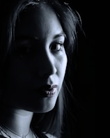 A close-up portrait of a person with dramatic lighting creating stark contrasts between light and shadow across the face. The image is predominantly in grayscale, with some visible texture on the skin. The person appears to be gazing directly at the camera, contributing to an intense and mysterious atmosphere.
