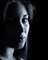 A close-up portrait of a person with dramatic lighting creating stark contrasts between light and shadow across the face. The image is predominantly in grayscale, with some visible texture on the skin. The person appears to be gazing directly at the camera, contributing to an intense and mysterious atmosphere.