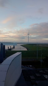 A large wind turbine stands near a body of water, surrounded by grassy land under a sky with scattered clouds. In the foreground, there is a modern building with a curved roof.