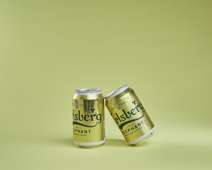 Two metallic cans with branding are positioned against a light green background. One can is slightly tilted and leaning against the other.
