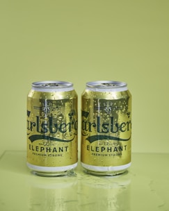 Two Carlsberg Elephant beer cans stand side by side on a flat surface, with visible condensation on their surfaces. The cans are predominantly gold with the Carlsberg logo and elephant imagery. The background is a solid, light green shade.