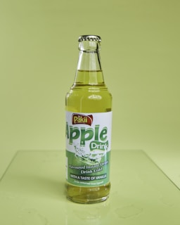 A bottle of Pakii Apple Drink sits on a light green surface, filled with a yellowish liquid. The bottle has a metal cap and a label that mentions it is a flavored instant drink with a taste of vanilla. The green label features water splash imagery and text highlighting the apple flavor.