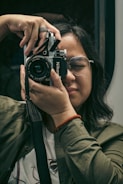 A person with glasses is holding up a vintage Nikon camera, preparing to take a photo. The individual is wearing a green jacket and a red bracelet, focused and with eyes gently closed. The background is blurred, emphasizing the act of photography.