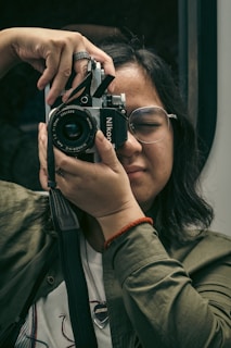 A person with glasses is holding up a vintage Nikon camera, preparing to take a photo. The individual is wearing a green jacket and a red bracelet, focused and with eyes gently closed. The background is blurred, emphasizing the act of photography.