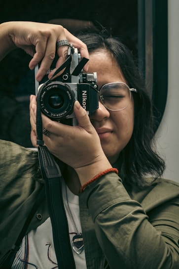 A person with glasses is holding up a vintage Nikon camera, preparing to take a photo. The individual is wearing a green jacket and a red bracelet, focused and with eyes gently closed. The background is blurred, emphasizing the act of photography.