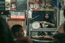 A woman wearing a hijab works in a food stall, packaging food items to sell. The stall is decorated with various menu signs and a neon sign displaying 'OPEN' and 'WELCOME'. The stall appears to be part of a busy market with a warm and inviting atmosphere.