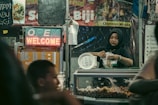 A woman wearing a hijab works in a food stall, packaging food items to sell. The stall is decorated with various menu signs and a neon sign displaying 'OPEN' and 'WELCOME'. The stall appears to be part of a busy market with a warm and inviting atmosphere.