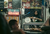 A woman wearing a hijab works in a food stall, packaging food items to sell. The stall is decorated with various menu signs and a neon sign displaying 'OPEN' and 'WELCOME'. The stall appears to be part of a busy market with a warm and inviting atmosphere.