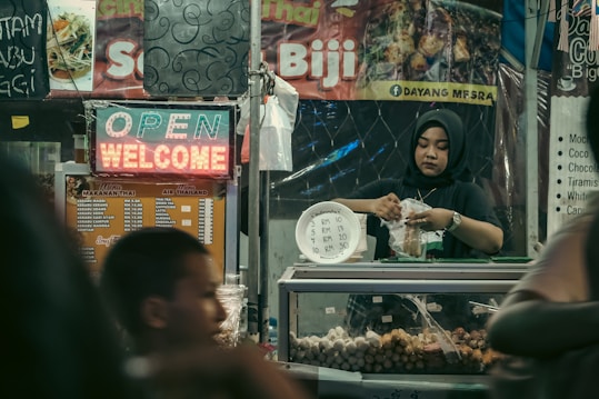 A woman wearing a hijab works in a food stall, packaging food items to sell. The stall is decorated with various menu signs and a neon sign displaying 'OPEN' and 'WELCOME'. The stall appears to be part of a busy market with a warm and inviting atmosphere.