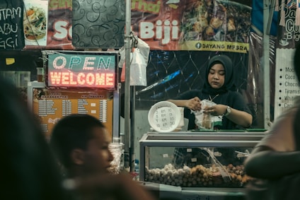 A woman wearing a hijab works in a food stall, packaging food items to sell. The stall is decorated with various menu signs and a neon sign displaying 'OPEN' and 'WELCOME'. The stall appears to be part of a busy market with a warm and inviting atmosphere.