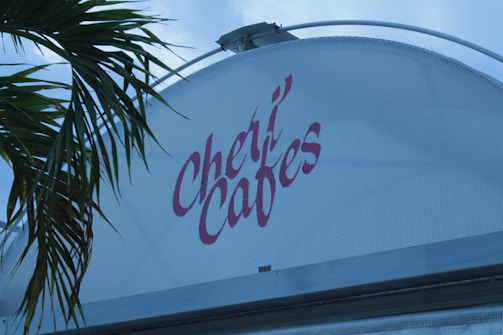 A building with a curved white facade features the stylized red lettering 'Cheri Cafes.' Palm tree fronds extend from the left side into the frame against a pale blue sky.